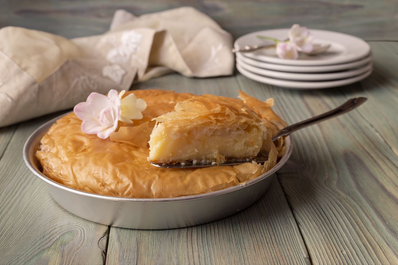 A slice of galaktoboureko, a Greek dessert, presented in a tie tin with white plates in the background
