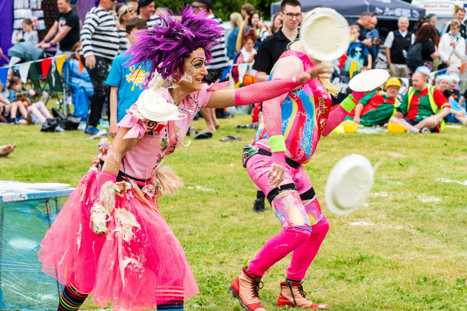 Coxheath, Maidstone. Woman in purple wig and pink costume, throwing a custard pie, with audience in the background watching, during the world custard pie Championship