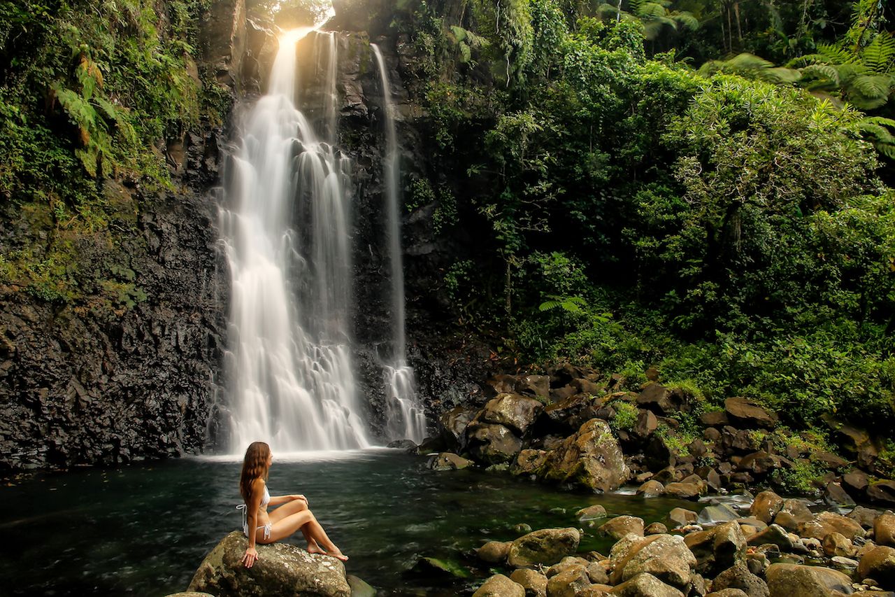 Young woman in bikini sitting by Middle Tavoro Waterfalls in Bouma National Heritage Park on Taveuni Island, Fiji