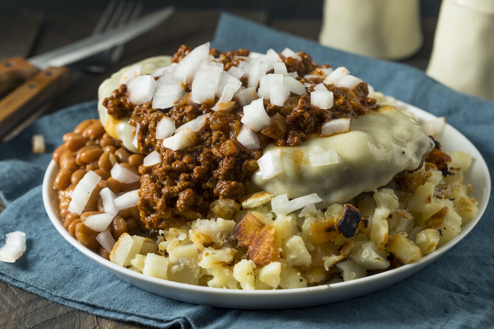 garbage plate with macaroni salad and beans topped with raw onions on a blue tablecloth