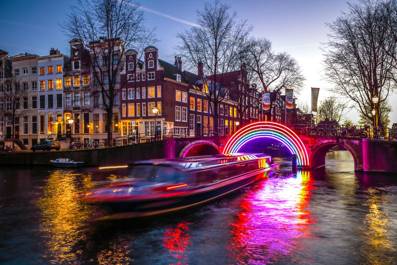A canal boat passing under a bridge with rainbow lights in Amsterdam