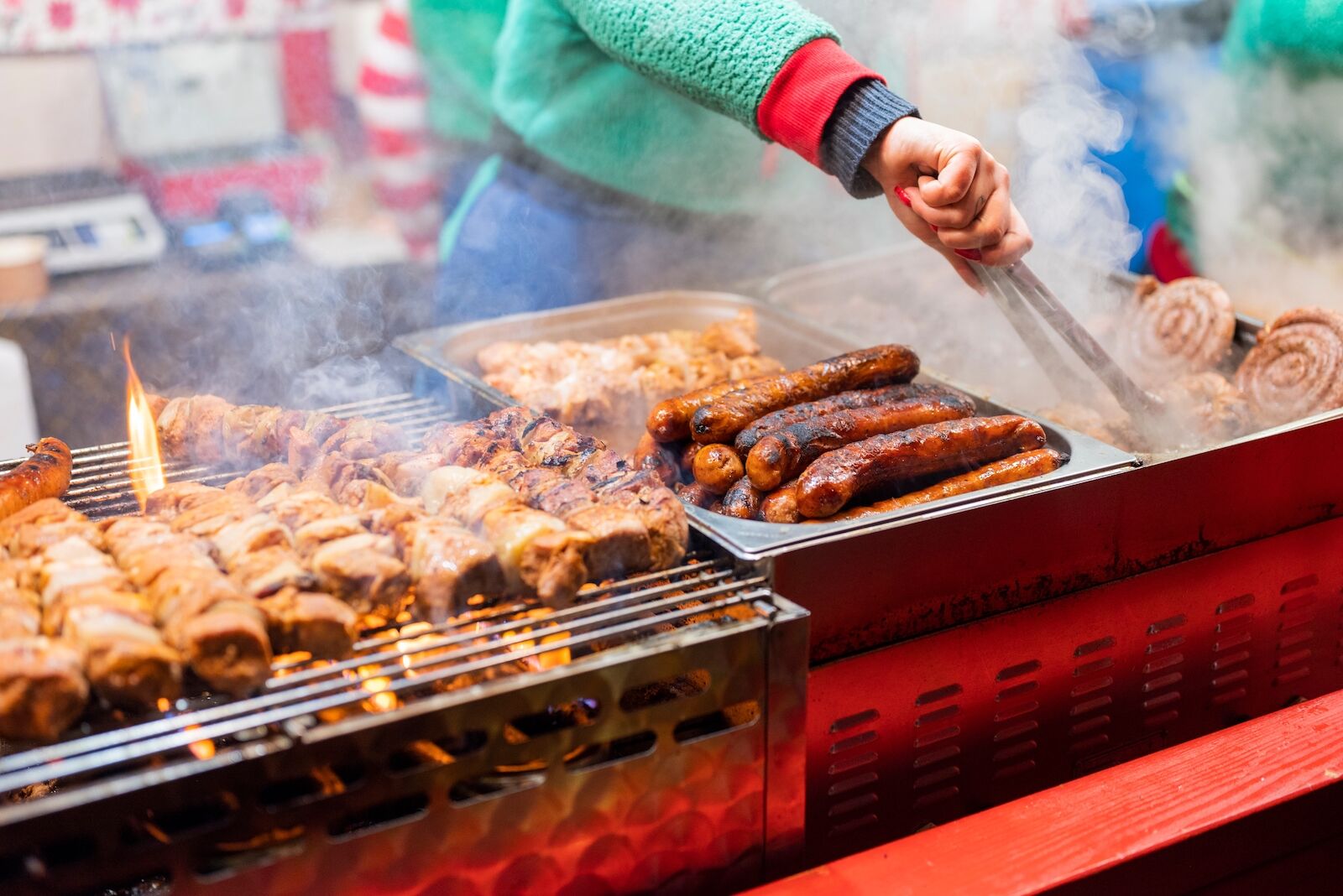 A woman with a pair of tongs in her hands grilling sausages and other meets at an outdoor food stall at the Gdansk Christmas markets, one of the best christmas markets in Europe