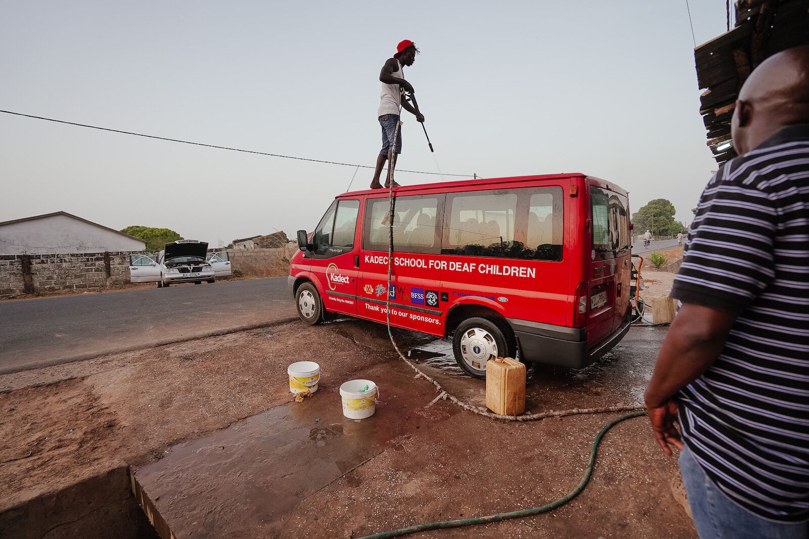 man standing on top of the bus from London to Gambia with a pressure washer to clean it up.