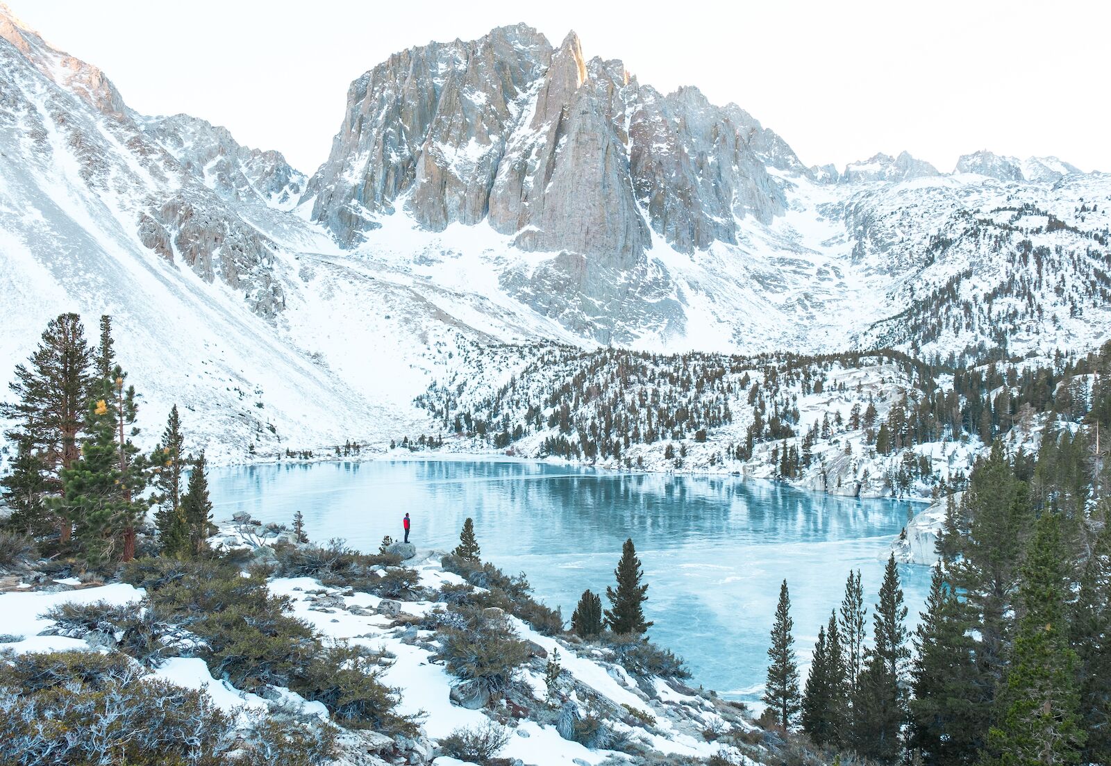 Man looks at a snowy peak bear a beautiful blue lake in the winter in Mammoth Lake California. Mammoth Lake is one of the best destinations for Spring Break 2022.