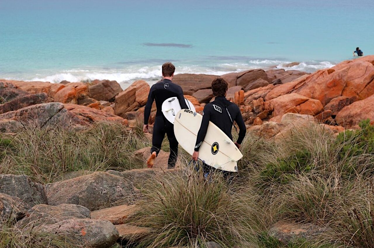 Two male surfers walk over rocks to the ocean in Perth Australia 