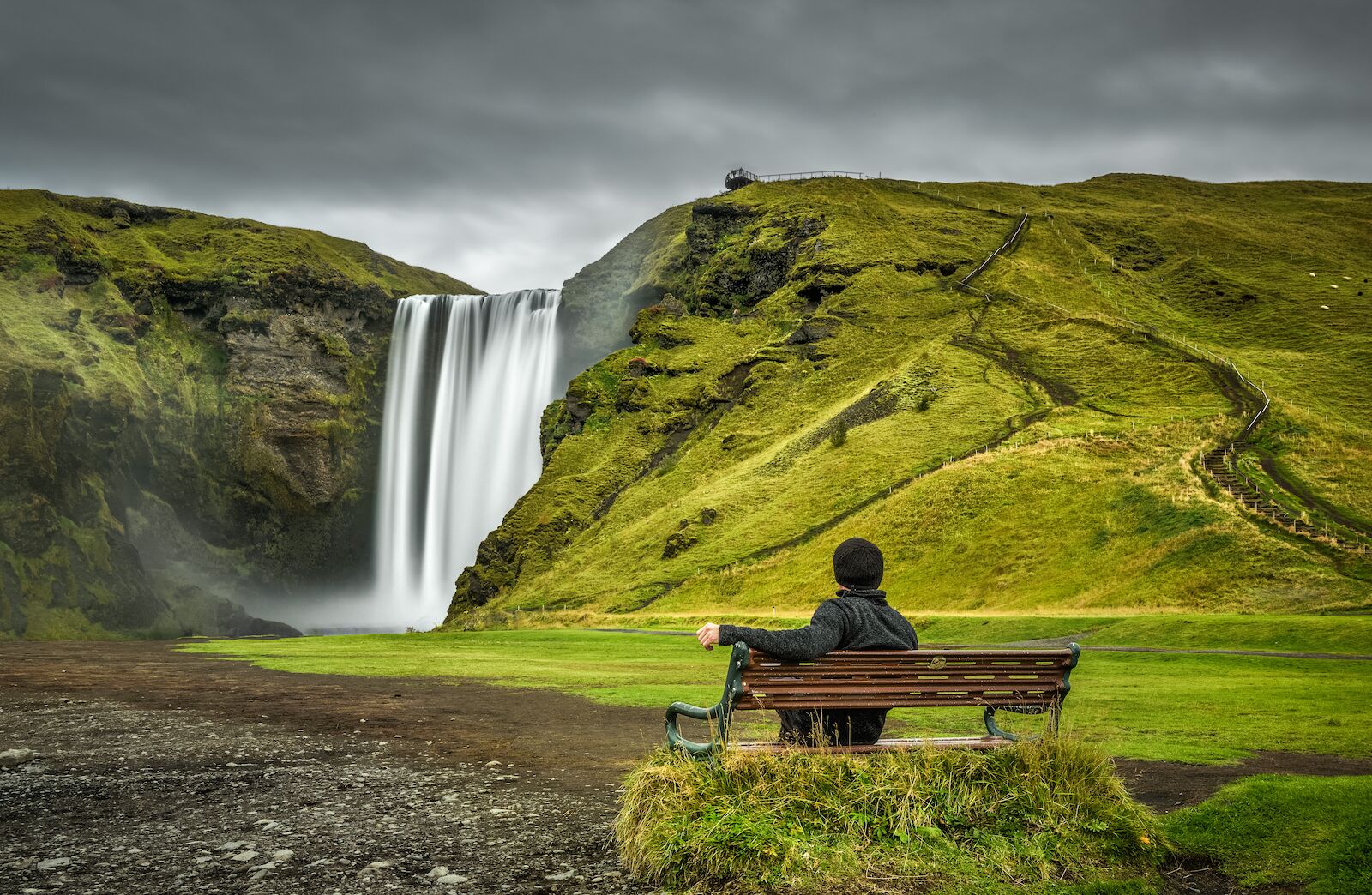 Person sitting in front of Skógafoss, the most famous waterfall in Iceland. In Icelandic, Skógafoss, is a composite word of skógar (forests) and foss (waterfall). 