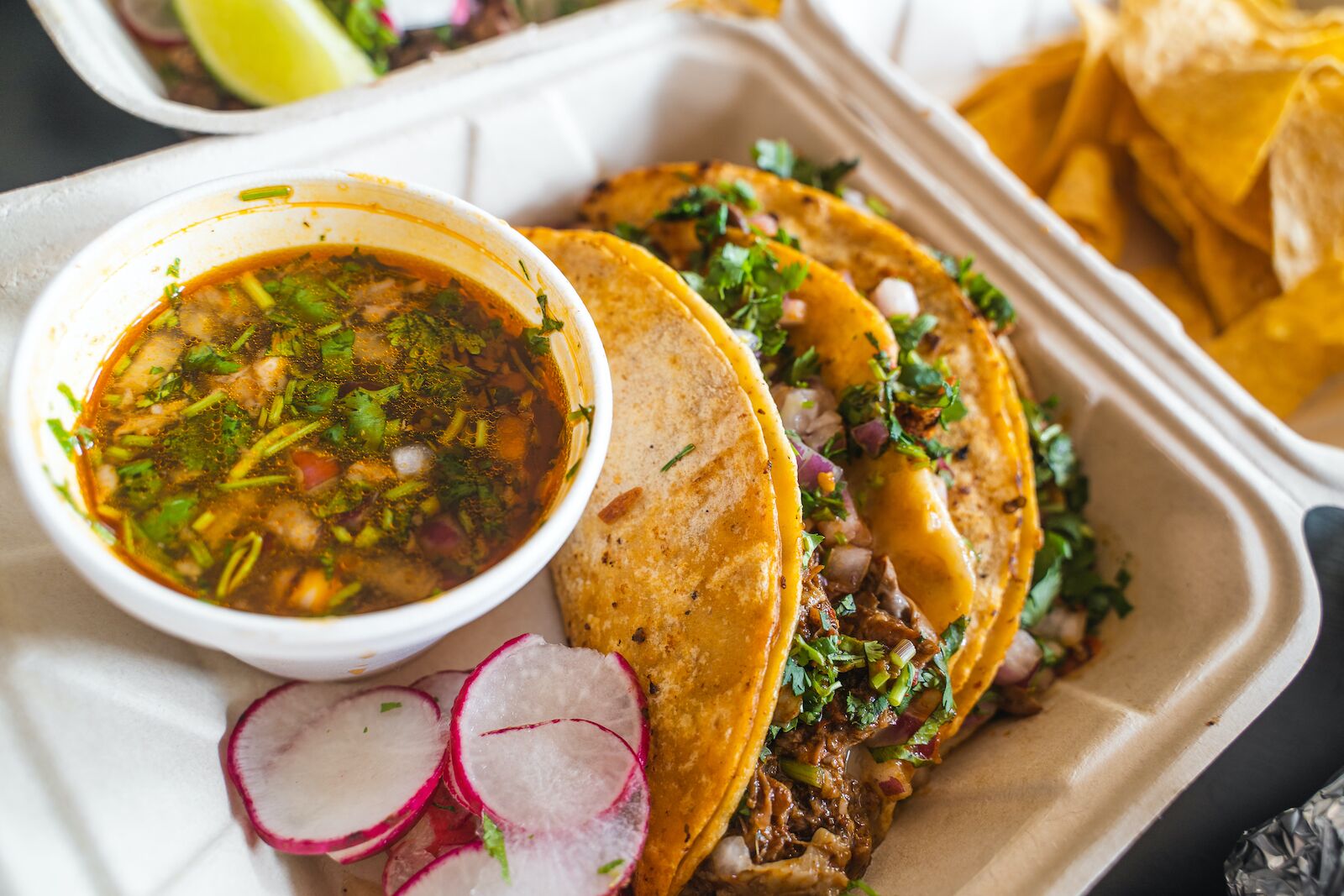 two birria tacos in a to-go container with a side of dipping sauce