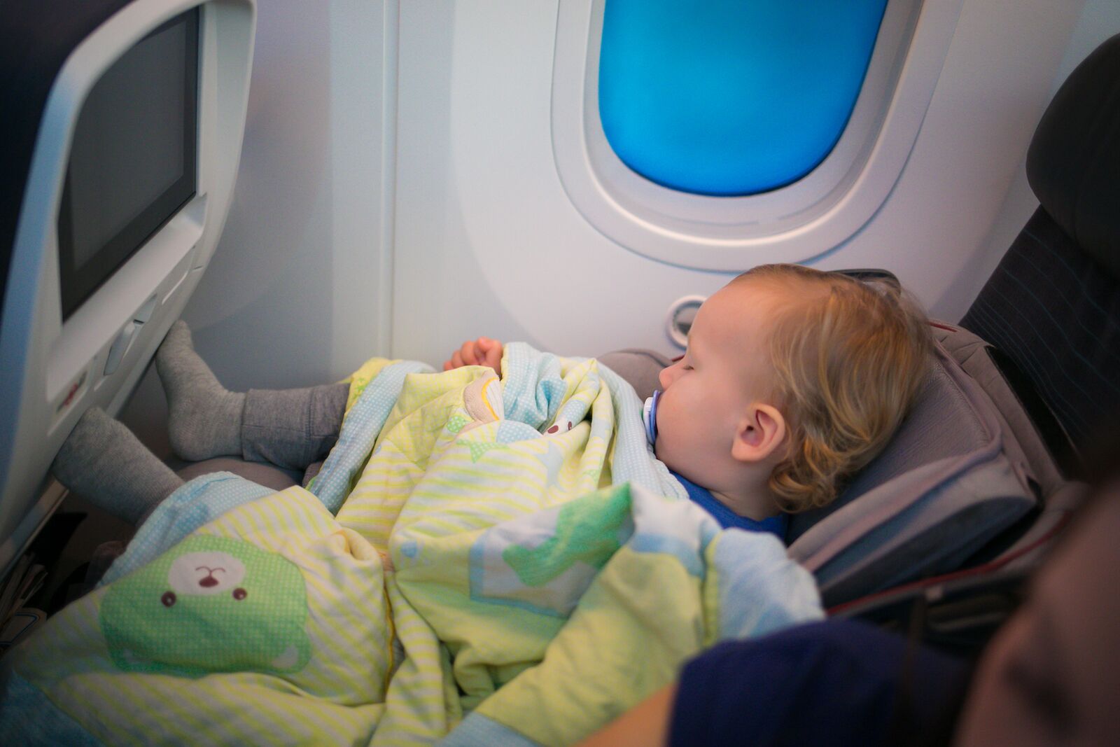 Flying with children: two year old baby sleeping in her own car seat setting on an ordinary seat on a commercial airliner. Concept photo of air travel with baby. Natural in-plane lightning conditions