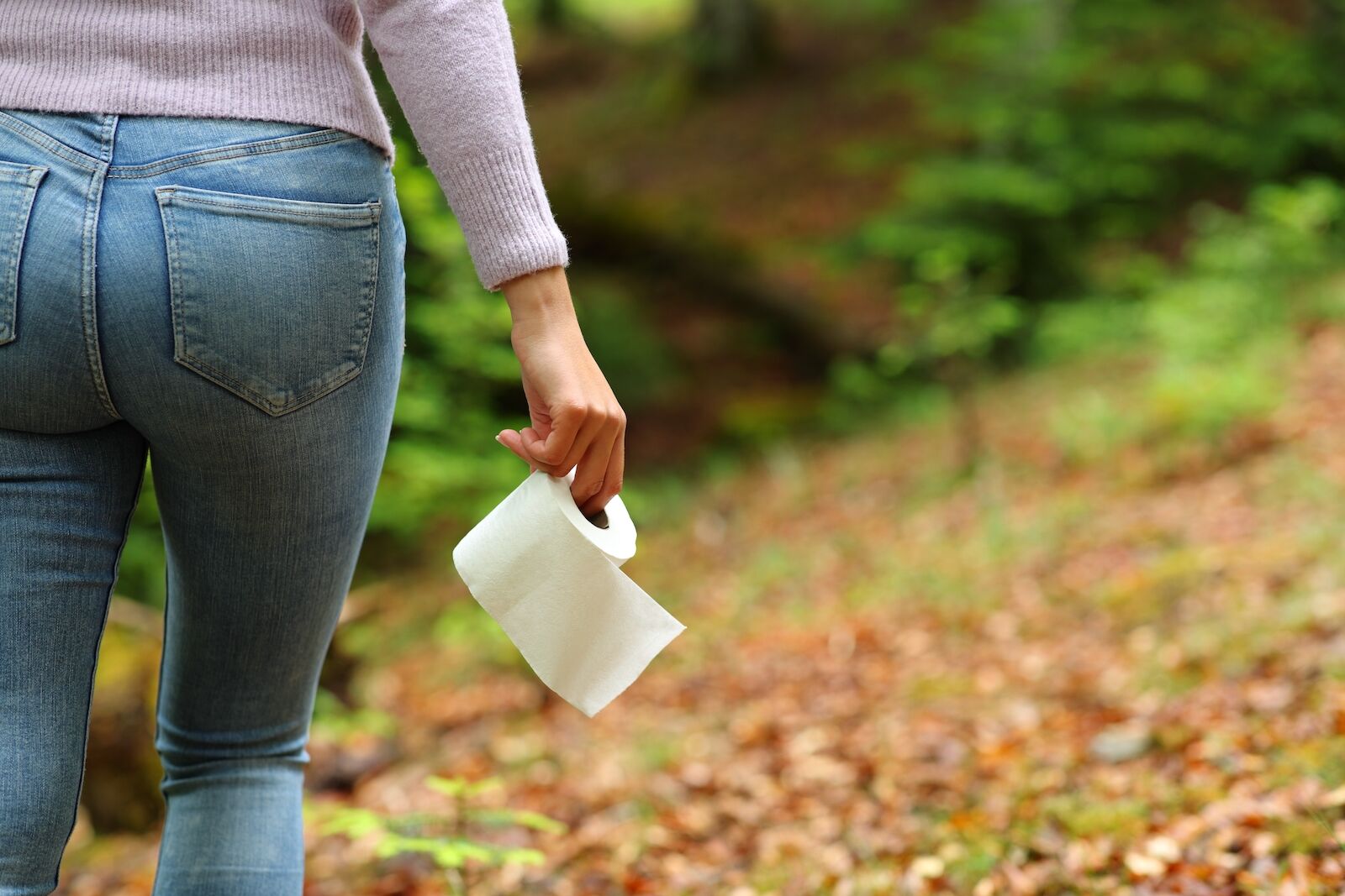view of woman wearing jeans from the back carrying roll of toilet paper in to the woods