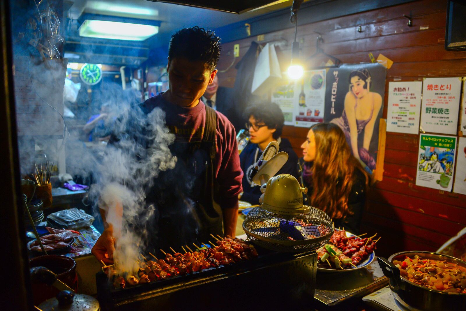 A man  grilled yakitori on Omoide Yokocho while diners sit at the counter 