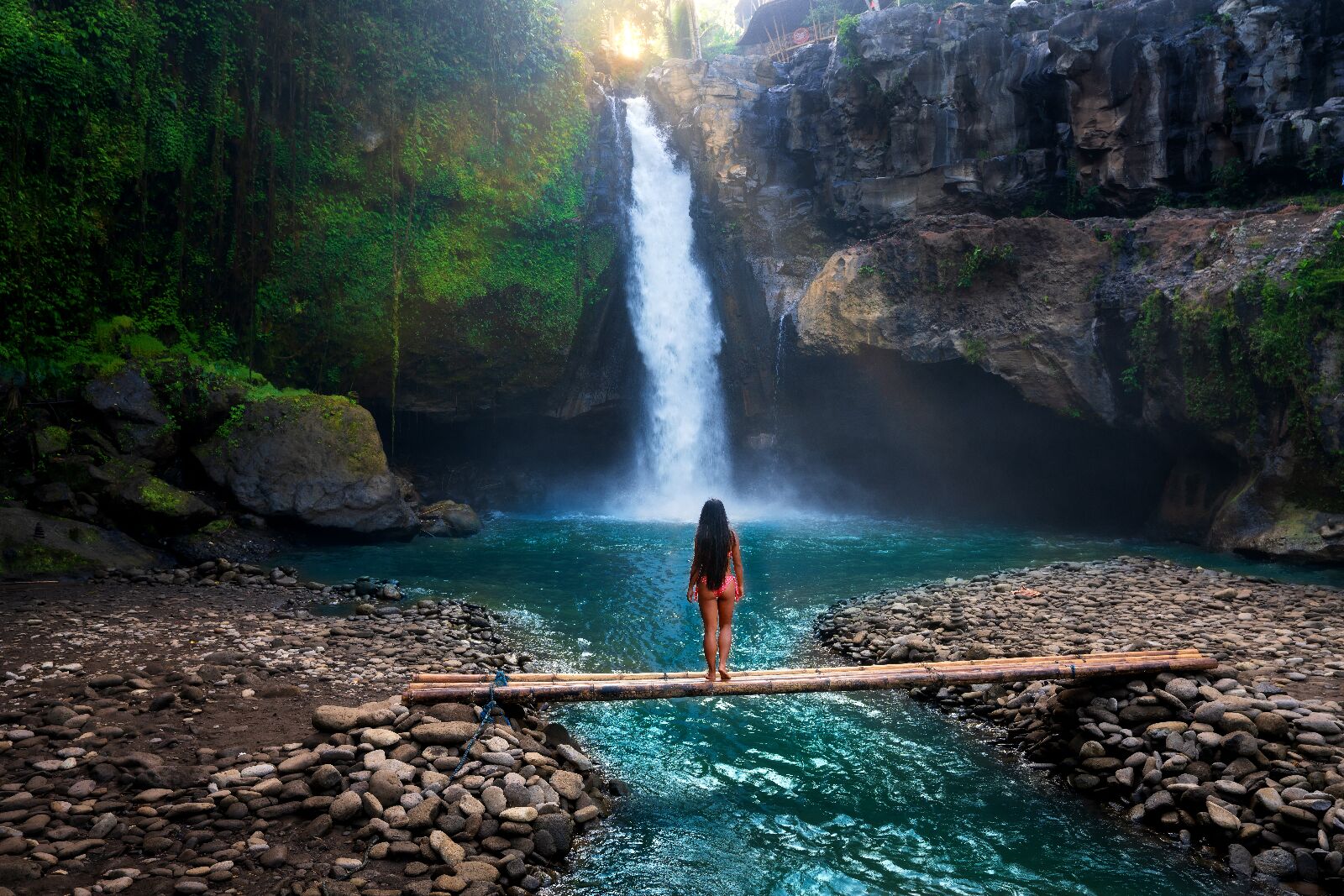 Rear view of young woman standing in front of waterfall with her hands raised. Female tourist with her arms outstretched looking at waterfall., bali on a budget