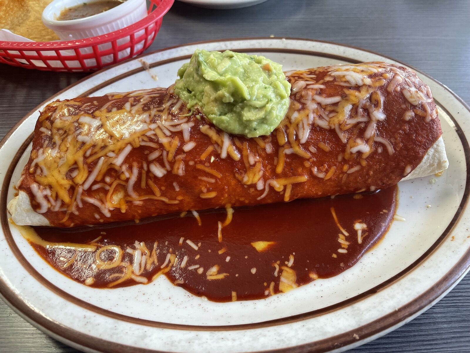 A large burrito topped with red sauce shredded cheese and guacamole on a white plate from La Cabanita