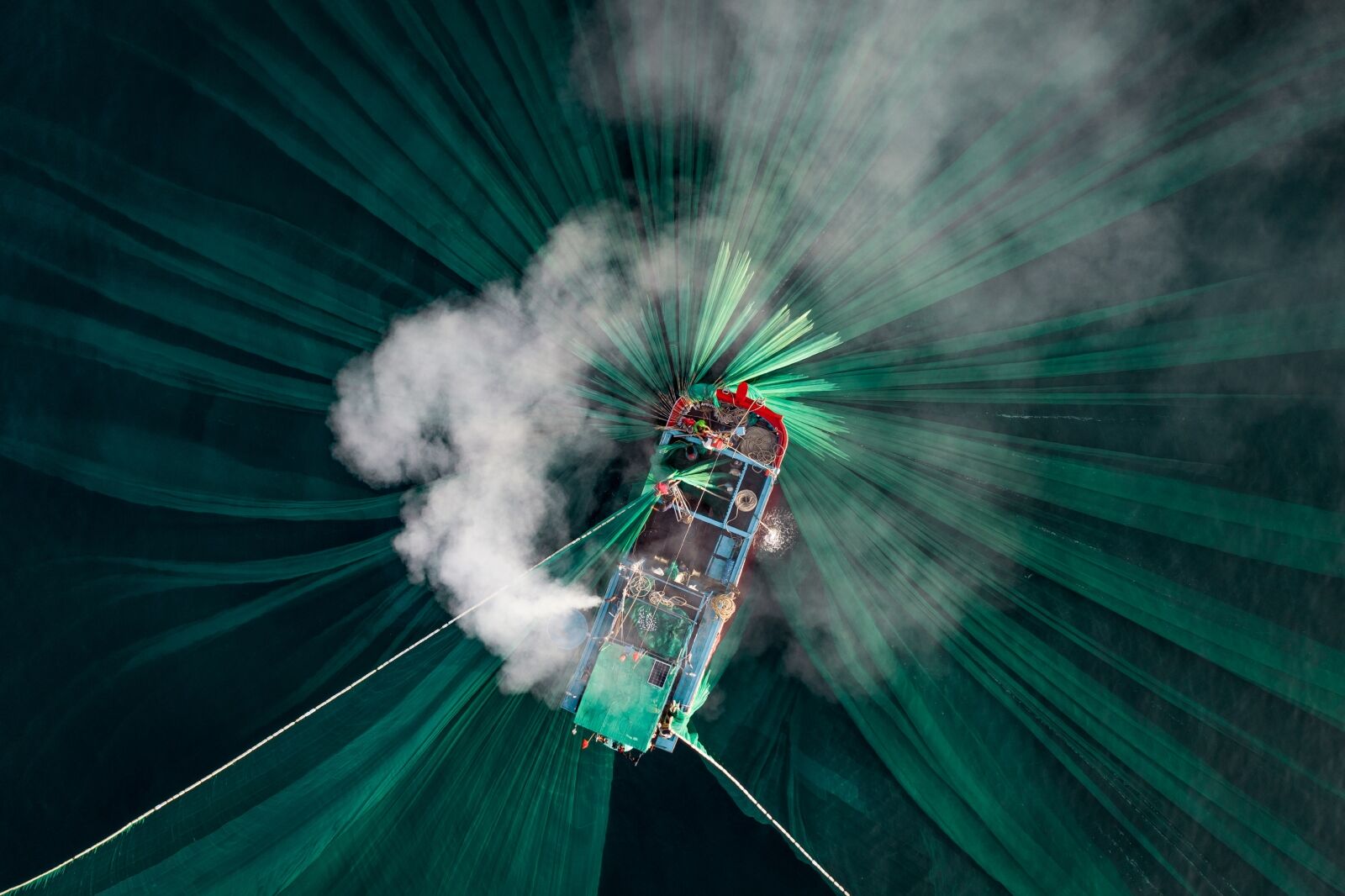 A fishing boat off Hon Yen as its long trail of smoke perfectly aligns with the shape of the green nets under the surface. An Ocean photographer of the year 2024 winning photo.