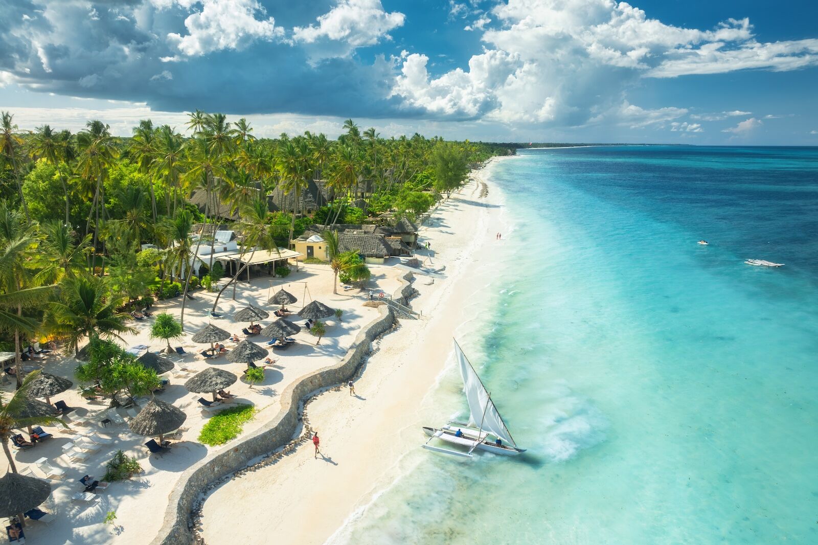 Aerial view of the sailboat on blue sea, empty white sandy beach, umbrellas at sunset. Summer in Zanzibar. Tropical scenery with boat, ocean with waves, green palms, sky. Top drone view. Exotic