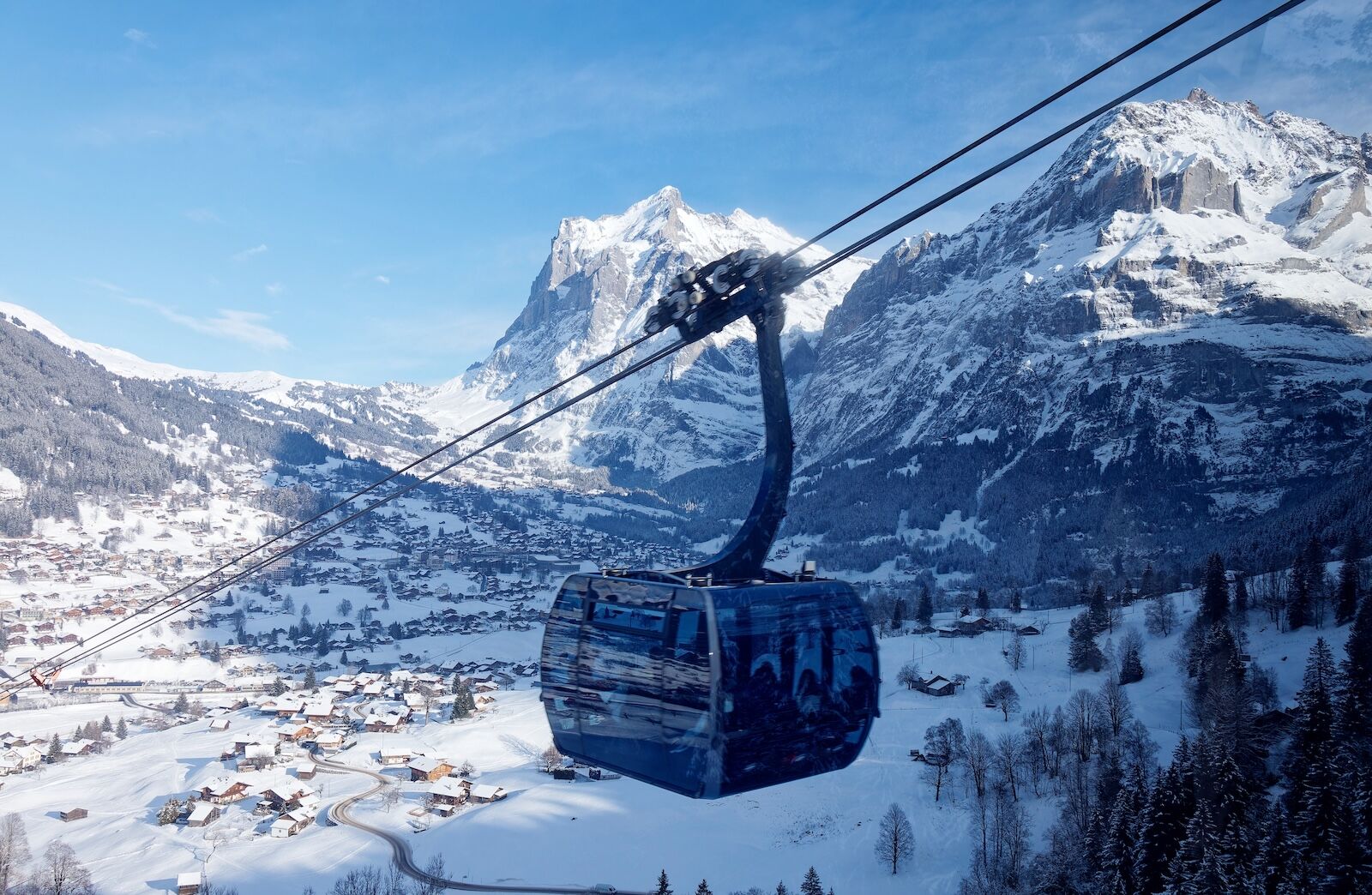 Winter scenery of the gondola of Eiger Express Cableway, which overlooks Grindelwald village on the snowy hillside and Wetterhorn mountain under blue sky in background, in Berner Oberland, Switzerland