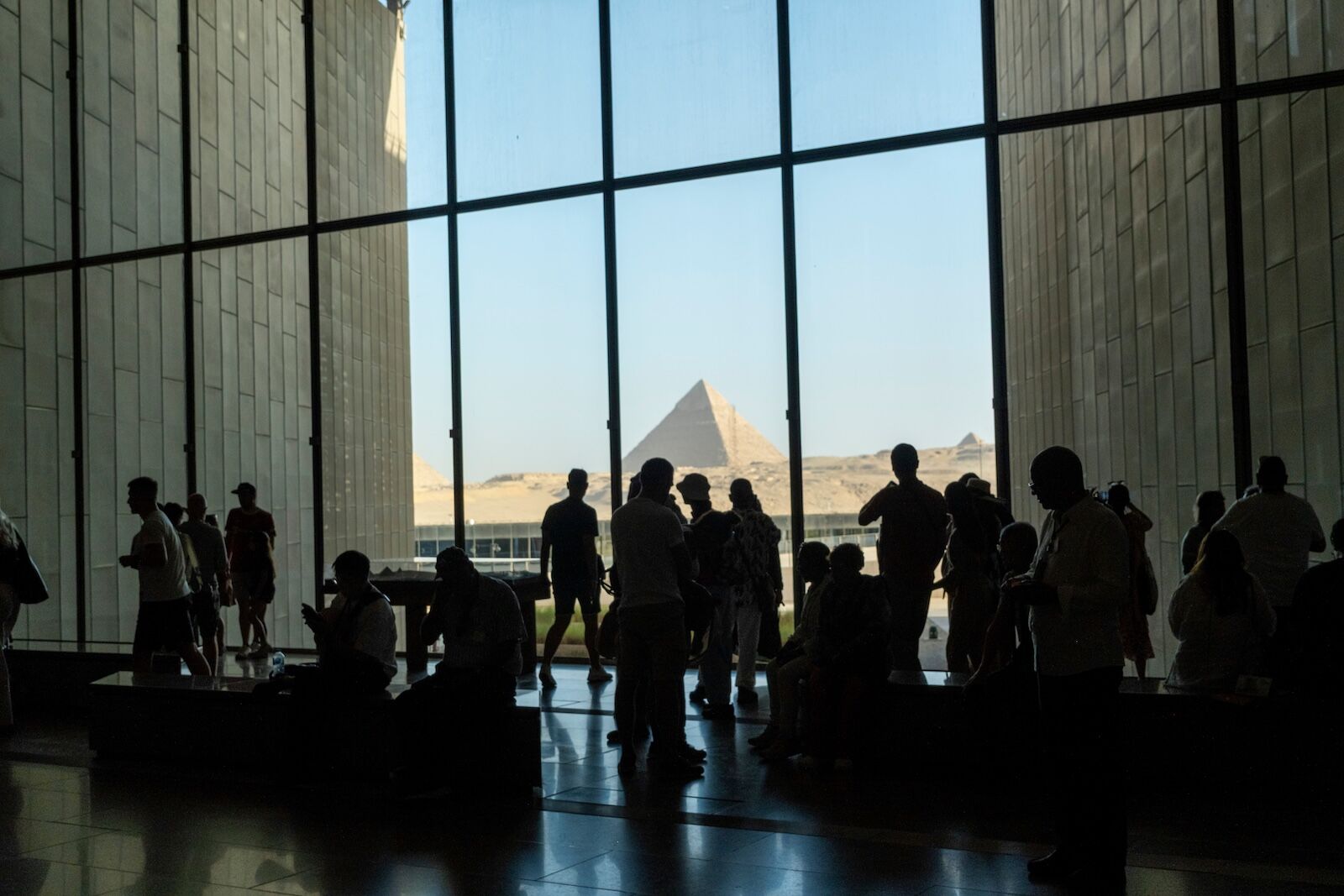Giza, Egypt. October 2, 2025: Grand Egyptian Museum (GEM). A large group of visitors are silhouetted against the museum's glass facade, with a pyramid of Giza clearly visible in the distance outside.