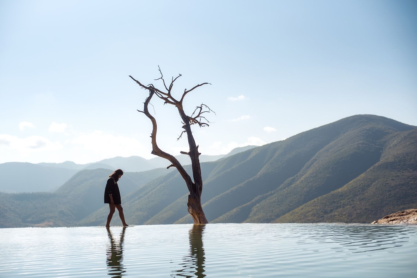Hierve El Agua, Oaxaca / Mexico - 01/04/2019: Female tourist walking on the edge of the fresh water springs in Hierve El Agua with beautiful mountains and ominous tree in the background
