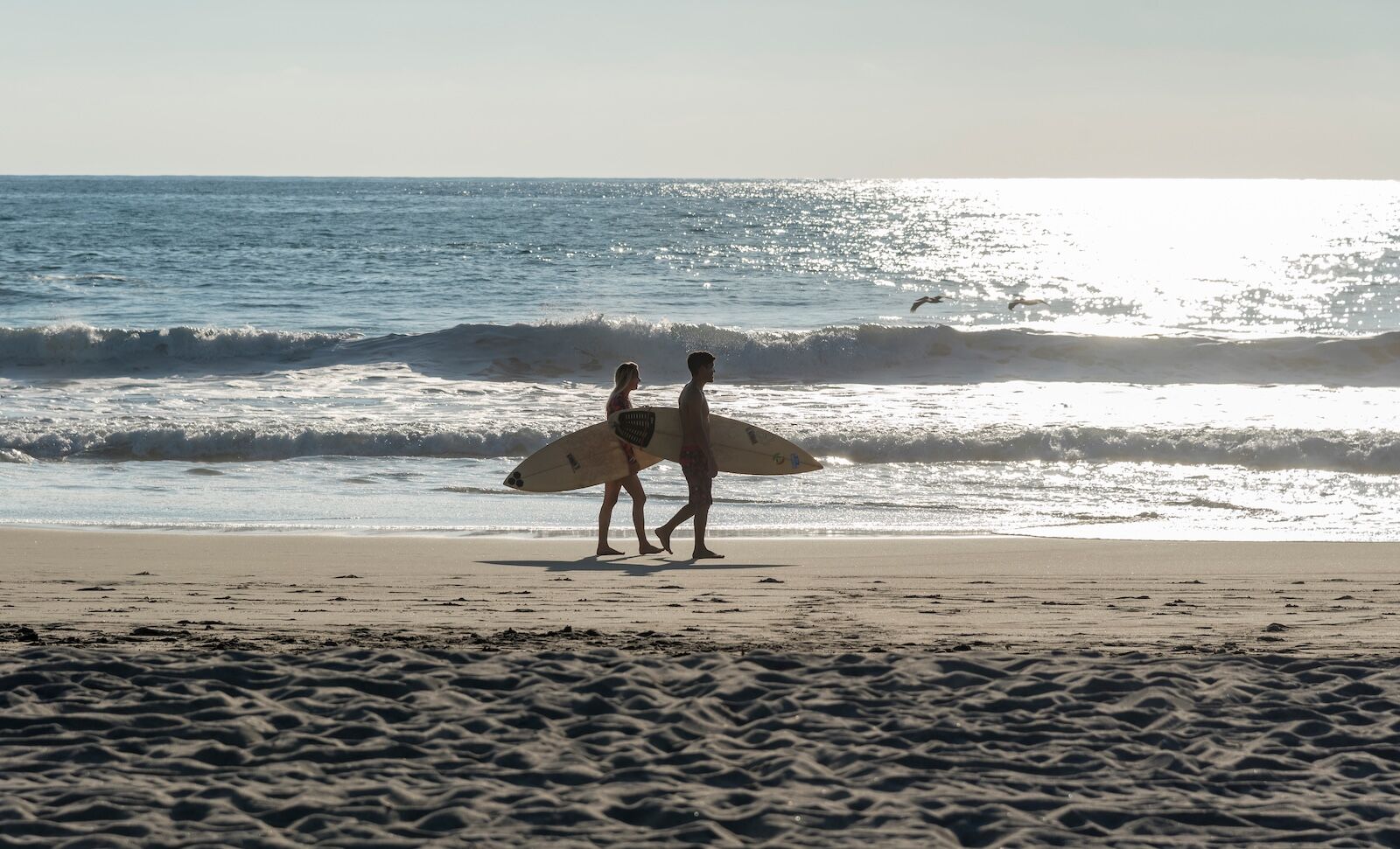 Mexico, Oaxaca, Puerto Escondido - November 10, 2019: a boy and a girl, a couple of surfers walking with surf on the beach, reflections of the sun at sea and pelicans flying
