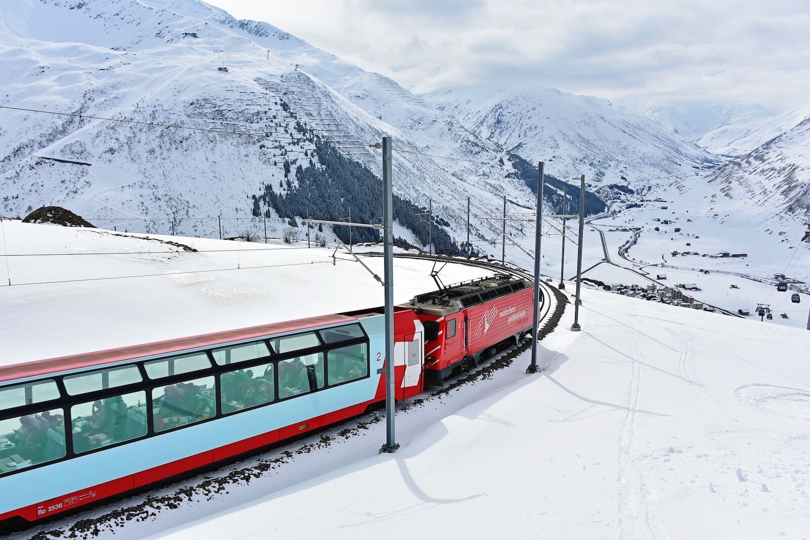 Andermatt Uri, Switzerland 03 February 2019: Glacier Express Switzerland, a red train in high mountains and Andermatt village, background snow and coluds in winter.